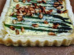 auf einem Brett liegt eine Quiche mit grünem Spargel und Pinienkernen