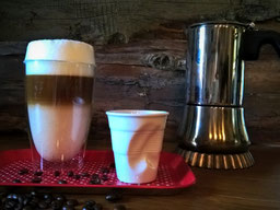 ein Latte Macchiato im Glas, eine zerbeulte Espressotasse aus Porzellan und ein Espressokocher