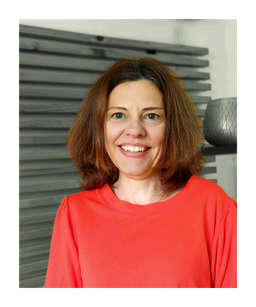 Foto von Sibylle Laabs: Sibylle steht in der Praxis (grauer Hintergrund) und lächelt in die Kamera, sie trägt ein rotes Oberteil