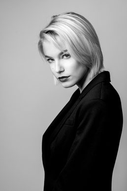 chwarzweiß-Portrait einer Frau im Fotostudio mit seitlicher Körperhaltung, direktem Blick in die Kamera und ruhigem, neutralem Hintergrund.