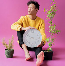  Person in gelben Pulli sitzt vor pinkem Hintergrund. Sie schaut zur Seite und hält eine Wanduhr in der Hand. Neben ihr stehen zwei Pflanzen.