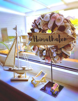 Segelboot, Liegestuhl und Muschelkranz mit Holzschild "Heimathafen" kombiniert zu einer maritimen Dekoration.