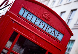 Oberer Teil einer roten britischen Telefonzelle, leicht schräg fotografiert. Man sieht die obere Ecke der Tür, darüber die Aufschrift "Telephone" und die Krone unter dem leicht gebogenen Dach. Im Hintergrund ein Haus mit vielen Fensterreihen.