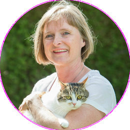 Lachende Astrid Huber in weißem T-Shirt hält dreifärbige Katze Nelly im Arm. Grünpflanzen im Hintergrund.