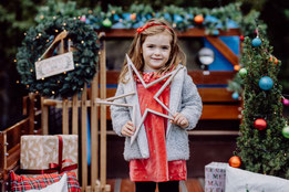 Kleines Mädchen hält einen Stern aus Holz in der Hand und steht vor bunter Weihnachtsdekoration