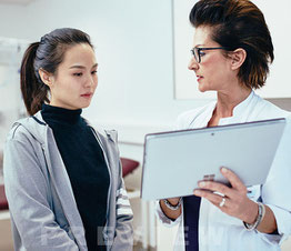 Ärztin in weißem Kittel erklärt einer Patientin am Tablet die nächsten Behandlungsschritte in einem modernen Behandlungsraum.