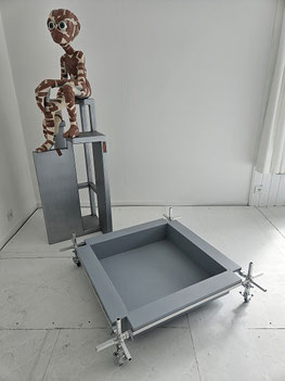 Sculptuur van een figuur volledig bedekt met pleisters zit op een verhoogde metalen structuur; ervoor staat een lage bak gemonteerd op een aluminium constructiesysteem met wielen.
