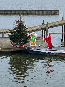 Auf einem Steg am Wasser steht ein weihnachtlich geschmückter Tannenbaum - daneben hockt der Frosch aus einem bekannten Märchen. Ein Weihnachtsmann auf einem Boot hat angelegt.