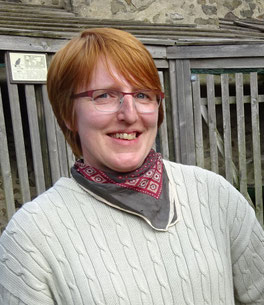 Foto von mir. Ich habe kurze rötliche Haare und eine Brille, trage einen hell khakifarbenen Baumwollpulli mit Zopfmuster und ein weinrot-beige-dunkelbraun gemustertes Halstuch. Im Hintergrund hölzerne Verschläge.
