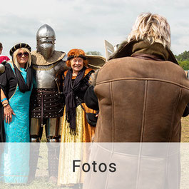 Im Vordergrund steht ein Mann in brauner Lederjacke, der mit seinem Smartphone ein Foto von einem Ritter und zwei älteren Damen in mittelalterlicher Gewandung macht.
