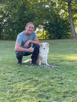 Wolfgang Raible kniet auf einer Wiese neben seinem sitzenden Hund Sammy, einem mittelgroßen, weiß-beigen Hund mit langem Fell, vor ihnen im Gras liegen zwei Leinen.