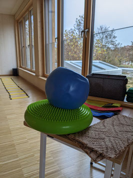 Ein Tisch mit Kleinen Sportgeräten steht vorne. Au dem Tisch liegen ein Ballkissen, ein ball, Therabänder und ein Teppich. Im Hintergrund liegt eine Koordinationsleiter. Große Fenster mit Blick nach außen.