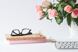 Auf einer weißen Tischplatte 2 Notizbücher, eine Brille, einige Klammern, eine helle Tastatur und in einer weißen Vase ein Strauß rosafarbener Rosen.