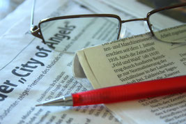 Nahaufnahme: Auf einer Zeitung liegen eine Brille und ein roter Bleistift mit Druckmine. 
