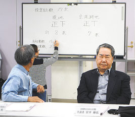 ホワイトボードに投票結果が記される。現地建て替えを主張してきた宮平委員(手前右)は目を閉じて結果を待つ=27日午後、市健康福祉センター