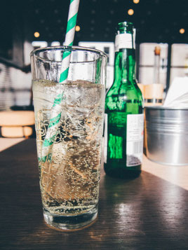 Glas mit farbloser Flüssigkeit mit Kohlensäure und Strohhalm, grüne Flasche im Hintergrund
