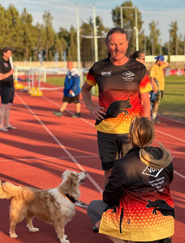 Wolfgang steht auf Laufbahn, trägt schwarz-rot-goldenes Sportshirt, spricht mit sitzender Frau in passendem Outfit, neben ihr steht Wolfgangs braun-weißer Hund Sammy.