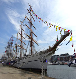 Ein eleganter weißer Großsegler, mit bunten Wimpeln über die Toppen geflaggt, liegt im Hafenbecken, dahinter sieht man die masten weiterer Großsegler. Am anderen Ufer sind Häuser und kleinere Boote zu erkennen. Blauer Himmel mit Schleierwolken.