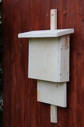 Fledermauskasten aus hellem Holz an einer Holzwand. Der Kasten istrechteckig, hell und hat ein abgesetztes Dach.