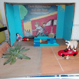 Gestalteter Schuhkarton zu einem Kinderbuch. Rechts ist ein Waschbärfigur auf einem roten Spielzeugauto. Links sitzt eine kleine Figur in der Ecke. Davor ist eine Pflanze zu sehen. 