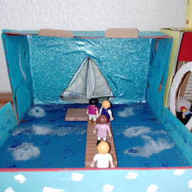 Es ist ein gestalteter Schuhkarton zu sehen. Playmobil Figuren stehen auf einem Steg und laufen über einen Steg auf ein Segelschiff im Hintergrund zu.