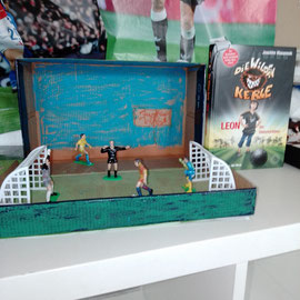Links ist das Buch "Die wilden Kerle" zu sehen. Rechts ist der gestaltete Schuhkarton dazu. Playmobilfiguren stehen zwischen zwei Toren auf einem Fußballplatz 