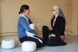 Eine Coachin sitzt mit ihrer Cochee auf Yogakissen und erklärt ihr eine energetische Methode, um ihre emotionalen Blockaden aufzulösen. 