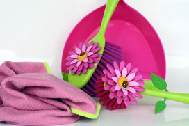 Ein Handfeger mit hellgrünem Griff und lila Borsten, mit einer künstlichen Blüte verziert, lehnt an einer aufgerichteten pinken Handschaufel. Davor ein rosa Putzlappen und eine rosa-grüne Spülbürste auch mit einer Blüte als Deko. 