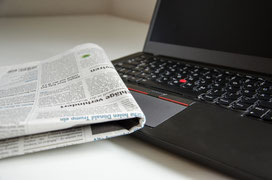 Auf einer weißen Tischoberfläche ein aufgeklappter schwarzer Laptop. Auf der linken vorderen Ecke seiner Tastatur liegt eine zusammengeklappte Zeitung.