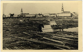 19th century photograph of logs piled up by the Daugava riverbank with Old Riga buildings in the background