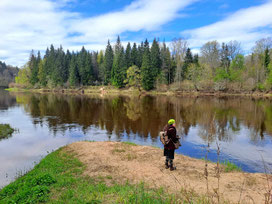 Girl with rucksack standing on bank on Gauja River with blue sky and forest background in Latvia