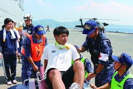 甲板から海上自衛官らが患者役を担架などで運んだ。「いせ」は2013年に台風30号被害の救援でフィリピン沖に派遣された。後方は患者を空輸した陸自ヘリ=2日、恩納村沖