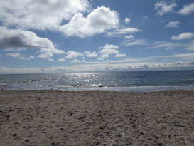 Hellbrauner Sandstrand vor blauem Meer. Am Himmel lugt die Sonne durch weiße Wolken