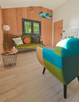 Cabinet de sophrologie de Gaëlle Pagès au Pertuis, Haute-Loire. Espace chaleureux en bois avec canapé vert et fauteuil coloré pour des séances de gestion du stress et burn-out dans un cadre serein et lumineux.