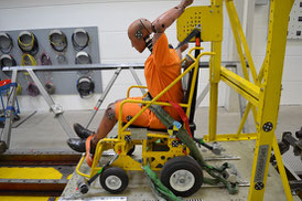 Crash-test dummy sits in a powered wheelchair on a sled. Wheelchair and dummy are secured with heavy duty restraint system.