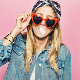 Julia, Bloggerin, Instagram Influencerin und zukünftige Autorin mit Tuch auf dem Kopf, Sonnenbrille und einer Kaugummiblase im Mund. der Hintergrund ist rosa und sie trägt eine blaue Jeansjacke