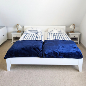 Man sieht ein gemütliches Doppelbett mit maritimer Bettwäsche und dunkelblauen Decken. Rechts und links des Bettes stehen Nachttische mit jeweils einer Lampe drauf