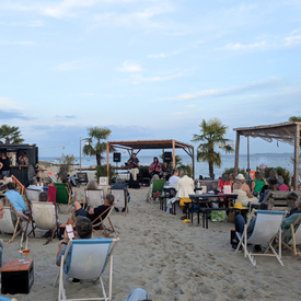 Eine Bühne am Strand. Die Band spielt Musik und drum herum stehen Liegestühle, in denen Menschen sitzen und der Musik lauschen.