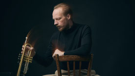 Trompeter Johannes Stange sitzend auf einem Stuhl, greift nach seiner Trompete