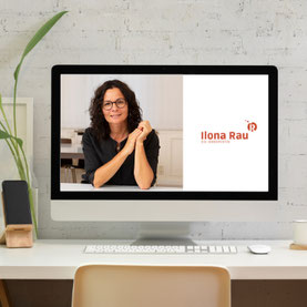 Schreibtisch auf dem ein iMac steht, auf dessen Bildschirm Dr. Ilona Rau zu sehen ist. Man sieht noch eine Pfllanze, eine Tasse, eine Computer-Maus und ein Smartphone auf einem Holzständer.