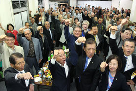桑江氏(前列右から3人目)のあいさつの後、ガンバロー三唱をする関係者ら=17日、コザ十字路前の事務所