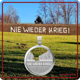Das Bild zeigt im Hintergrund die Waldbröer Mauer mit der Aufschrift Nie Wieder Krieg und im Vordergrund den silbernen Waldbröl Kettenanhänger, der die Mauer mit ebendieser Aufschrift zeigt