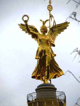 Sehenswürdigkeiten Berlin: Die Siegesgöttin Viktoria steht auf der Siegessäule, in der linken Hand hält sie einen Lorbeerkranz, in der rechten einen Stab. Auf dem Kopf schlägt ein Adler seine Flügel.