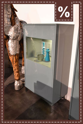 Hellgraue Vitrine mit Glasfront und Beleuchtung. Türkisfarbene Vasen innen. Prozentzeichen.