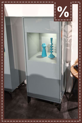 Hohe, hellgraue Vitrine mit Glasfront und Beleuchtung. Türkisfarbene Vasen innen. Prozentzeichen.