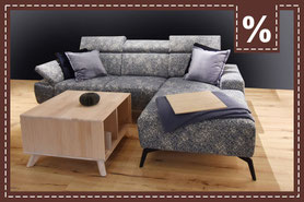 Graublau gemustertes L-Sofa mit Couchtisch aus Holz auf einem Holzboden. Prozentzeichen.