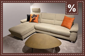 Beigefarbenes Ecksofa mit verstellbarer Kopfstütze, orangenen Kissen und Couchtisch. Prozentzeichen.