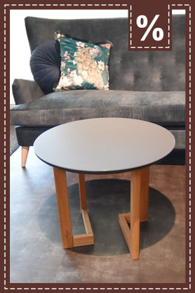 Runder grauer Couchtisch mit Holzbeinen vor einem dunkelgrauen Sofa. Prozentzeichen in der Ecke.