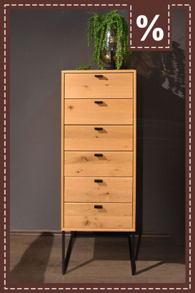 Eine hohe, schmale Kommode aus hellem Holz an einer dunklen Wand. Prozentzeichen in der Ecke.