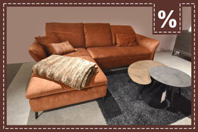 Rostbraunes L-Sofa mit Decke und Kissen, zwei runde Couchtische auf einem grauen Teppich. Prozentzeichen.